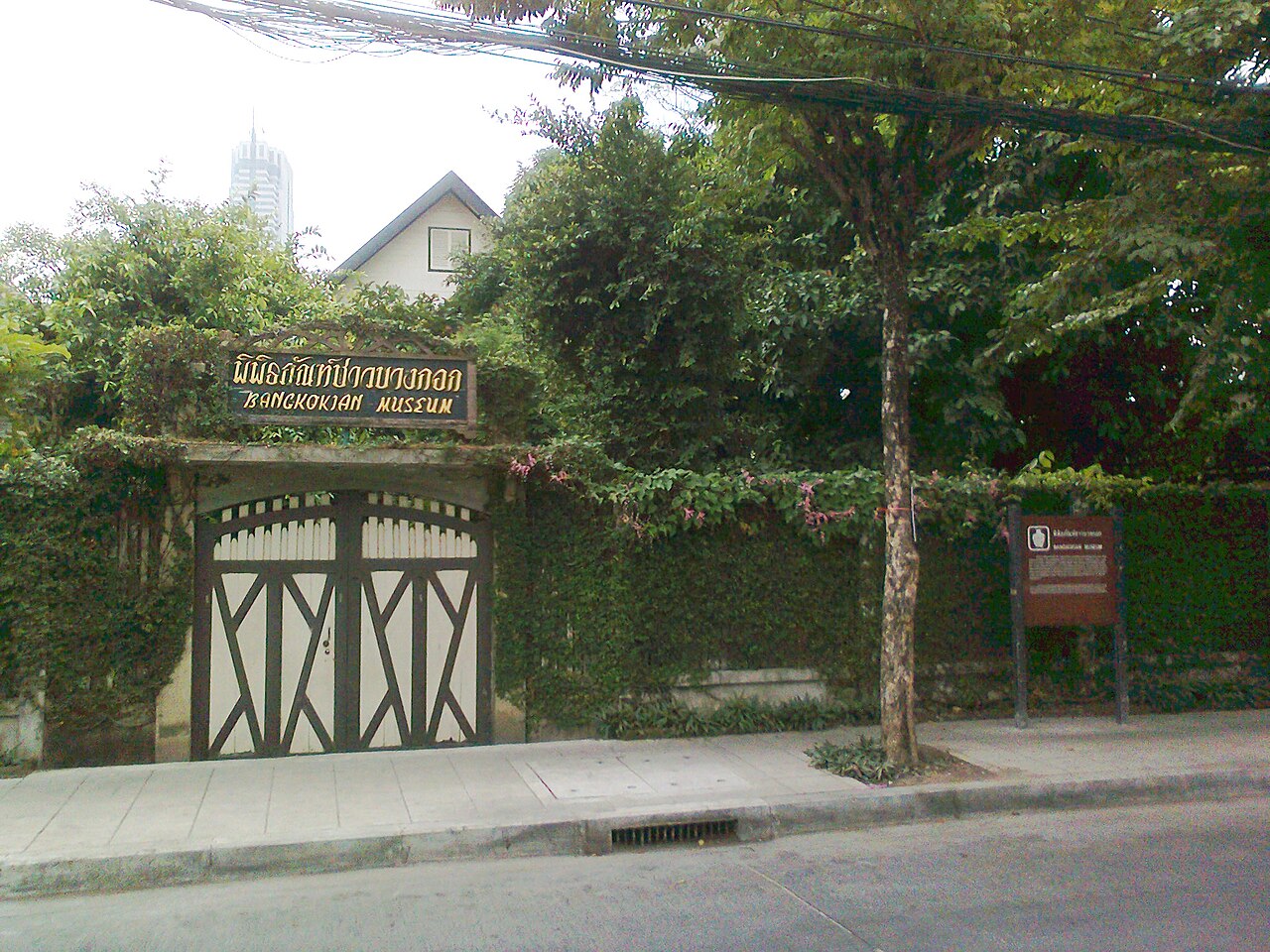 曼谷民俗博物館入口的鐵門與館名招牌，門內可見庭園